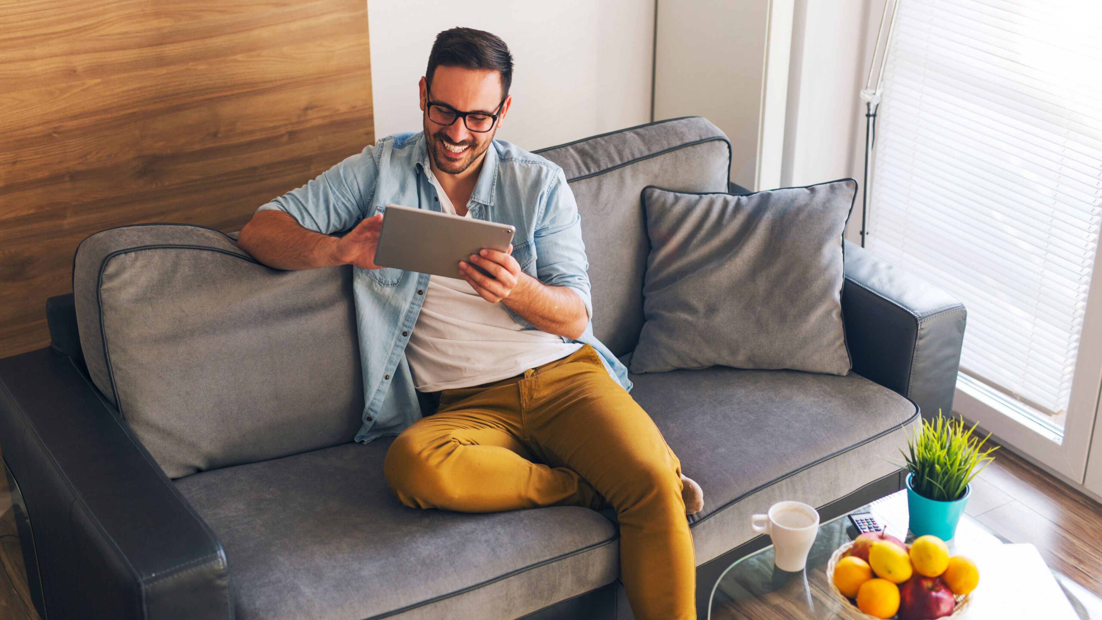 A smiling man in a button-down shirt and glasses sits on a sofa, looking at a tablet.